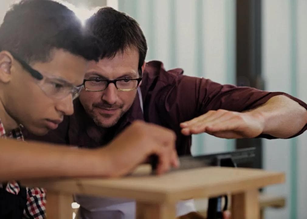 Two people: one appears to be a teacher, and the other is a student or apprentice. The teacher, a man wearing glasses and a maroon shirt, is intently observing the student's work. The student is looking at the wooden structure.