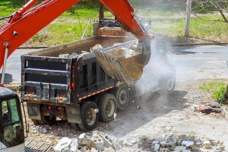 Excavator scooping crushed concrete into the back of a large truck which is on its way to landfill