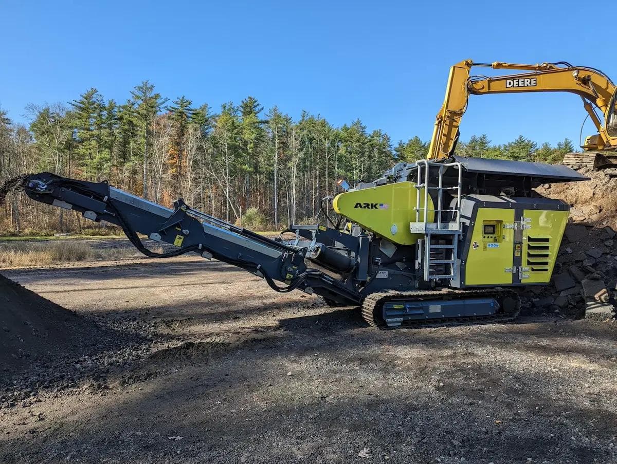 yellow jaw crusher being fed by a john deere excavator with sphalt or blacktop driveway. The asphalt is being crushed down into a small size and is being stockpiled on the other side at the end of the conveyor
