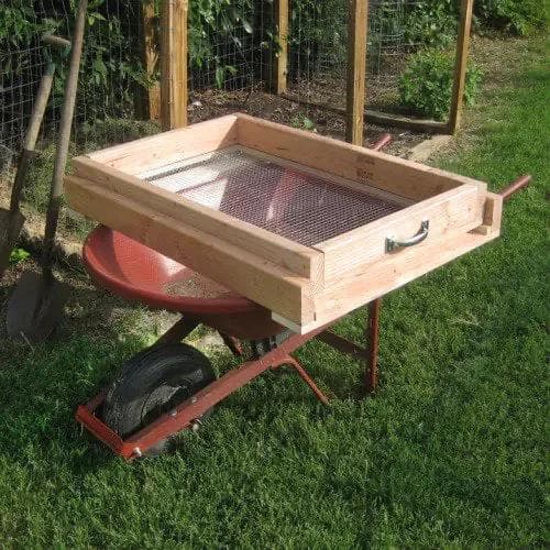 wooden and metal compost sifter with handle on top of a brown wheelbarrow in a garden