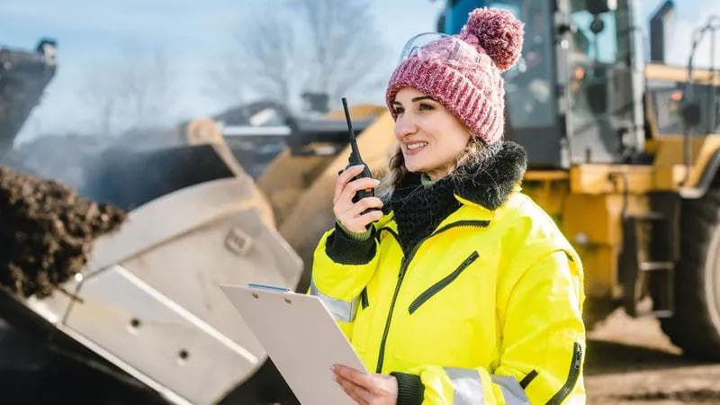 Worker in high-visibility yellow jacket and pink winter hat holding a clipboard and walkie-talkie, with construction equipment in the background