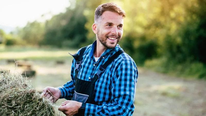 man in a blue and black checked shirt on a farm, he is holding a large bail of hay